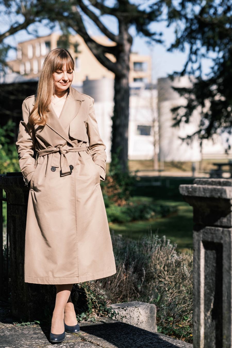 Une femme en trench beige, les mains dans les poches, dans un jardin
