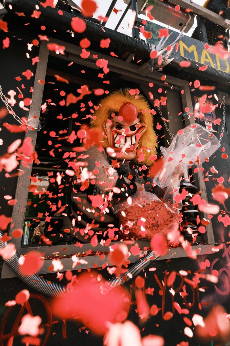 Un personnage masqué de carnaval lance une pluie de confettis rouges depuis un char